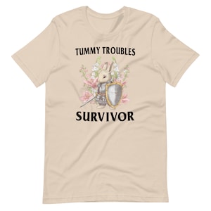 May include: Beige t-shirt featuring a cartoon bunny in armour holding a sword. The text "Tummy Troubles Survivor" is printed on the shirt.