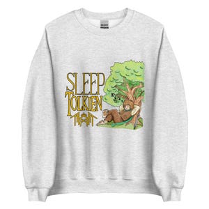 Peut inclure: Un sweat-shirt gris clair à col rond avec une impression graphique représentant un personnage dormant sous un arbre. Le texte "SLEEP TOLKIEN" est imprimé en jaune avec un contour noir à côté de l'illustration, incluant une épée et du feuillage vert.