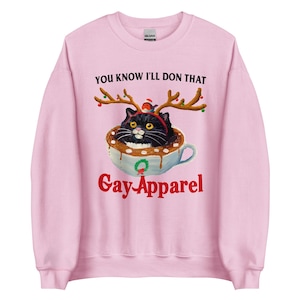 Puede incluir: Sudadera rosa claro con un gato negro con cuernos de reno en una taza de chocolate caliente. El texto "YOU KNOW I'LL DON THAT" está encima del gato y "Gay Apparel" debajo. Diseño festivo.