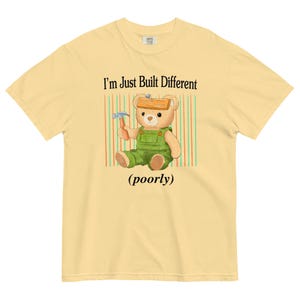 May include: A yellow t-shirt with a cartoon bear wearing a green jumpsuit and a wooden board on its head. The bear is holding a hammer. The text on the shirt reads "I'm Just Built Different (poorly)"