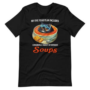 May include: Black t-shirt with a graphic of a black cat wearing blue goggles in a bowl of soup. The text reads "MY FIVE YEAR PLAN INCLUDES CONSUMING A VARIETY OF DIFFERENT SOUPS."