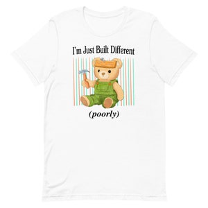 Könnte beinhalten: Weißes T-Shirt mit einer Cartoon-Illustration eines Teddybären, der eine Latzhose trägt und einen Hammer hält. Der Bär hat ein Stück Holz auf dem Kopf. Der Text "I'm Just Built Different (poorly)" ist auf das T-Shirt gedruckt.