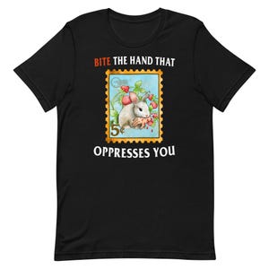 Peut inclure: T-shirt noir avec un graphique blanc d'un lapin mordant une main. Le graphique est dans le style d'un timbre-poste vintage avec le texte "Bite the hand that oppresses you".