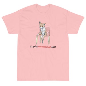 May include: A pink t-shirt with a cartoon fox sitting on a chair. The text "it's giving existential dread, bestie" is printed below the fox.