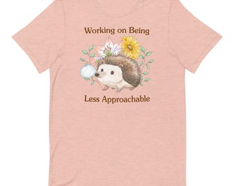 Puede incluir: Una camiseta rosa claro con un erizo marrón soplando una burbuja. El erizo está rodeado de flores y hojas. El texto "Working on Being Less Approachable" está impreso en la camiseta.