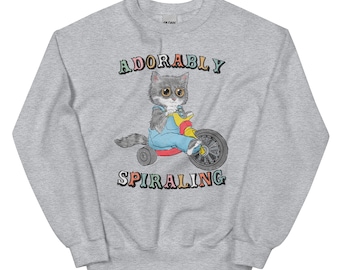 Peut inclure: Un sweat-shirt gris avec un chat de dessin animé conduisant un tricycle. Le texte "ADORABLY SPIRALING" est imprimé sur le sweat-shirt en couleurs arc-en-ciel.