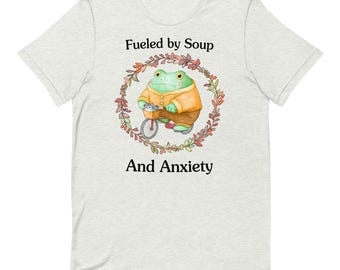 Puede incluir: Una camiseta gris claro con una rana verde montando una bicicleta en una corona de hojas de otoño. El texto "Fueled by Soup and Anxiety" está impreso en la camiseta.