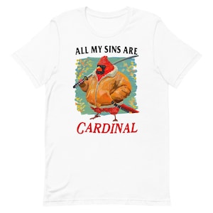 May include: White t-shirt featuring a red cardinal wearing a brown jacket and holding a sword. The text "All my sins are cardinal" is printed on the shirt.