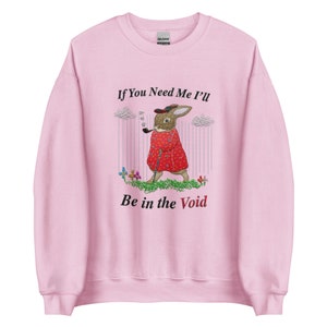 May include: A light pink sweatshirt with the text "If You Need Me I'll Be in the Void." The graphic features a rabbit in a red coat, hat, and smoking a pipe. The rabbit is standing in a field of grass and flowers.
