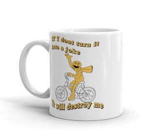 Può includere: Tazza in ceramica bianca con un'illustrazione gialla e nera di un burattino che guida una bicicletta. Il testo "Se non lo trasformo in uno scherzo, mi distruggerà" è stampato sulla tazza.