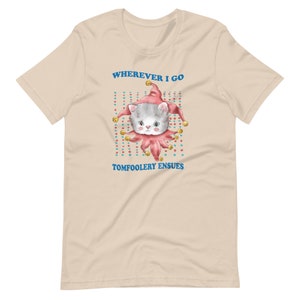 Op de afbeelding: Een beige T-shirt met een cartoonkat die een narrenmuts draagt. De tekst "WHEREVER I GO TOMFOOLERY ENSUES" staat op het shirt gedrukt.