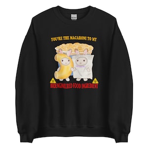 Könnte beinhalten: Schwarzer Pullover mit einer weißen Grafik von zwei Cartoon-Kaninchen, die als Makkaroni und Käse verkleidet sind. Der Text "You're the macaroni to my" und "Bioengineered Food Ingredient" ist in Rot gedruckt.