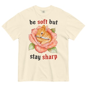 Könnte beinhalten: Cremefarbenes T-Shirt mit einer Aquarellillustration eines Kaninchens, das ein Messer in einer Rose hält. Der Text "be soft but stay sharp" ist oberhalb und unterhalb des Bildes in gotischer Schrift gedruckt.