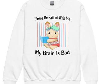May include: White sweatshirt with the text "Please Be Patient With Me" and "My Brain Is Bad". The graphic features a cartoon rabbit reading a book, wearing a green book on its head, and a red dress.