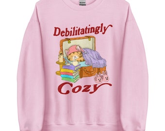 Può includere: Felpa rosa chiaro con le parole "Debilitatingly Cozy" in corsivo rosso. La grafica presenta un gatto in una valigia con una coperta, libri e pantofole a forma di coniglietto. Una candela e una tazza di tè sono anch'esse nell'illustrazione.