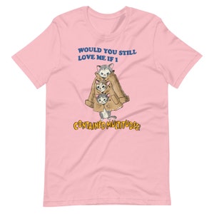Op de afbeelding: Roze T-shirt met een afbeelding van drie katten die een bruine jas dragen. De tekst op het shirt luidt "Would you still love me if I contained multitudes?"