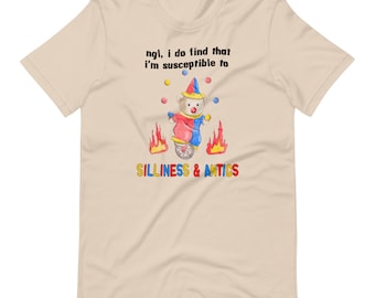 May include: A beige t-shirt with a cartoon illustration of a teddy bear dressed as a clown riding a unicycle. The bear is juggling balls and there are flames on either side of the unicycle. The text "ngl, i do find that i'm susceptible to silliness & antics" is printed on the shirt.