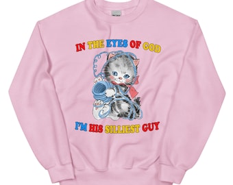 Pode incluir: Camisola rosa com um gato de desenho animado segurando uma bola de lã azul. O texto "In the eyes of God I'm his silliest guy" está impresso em letras coloridas na camisola.