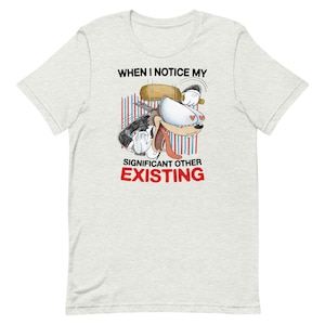 Puede incluir: Camiseta gris claro con un perro de dibujos animados con una expresión triste y el texto "WHEN I NOTICE MY SIGNIFICANT OTHER EXISTING" en negro y rojo. El perro tiene un gran ojo blanco con corazones.
