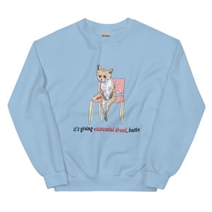 May include: Light blue sweatshirt featuring a watercolor illustration of a dog sitting on a pink chair. The text "it's giving existential dread, bestie" is printed in red below the image. The sweatshirt is made of a soft material.