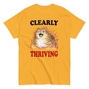Op de afbeelding: Een geel T-shirt met een cartoonkat in vlammen en de tekst "CLEARLY THRIVING" in zwart en rood.