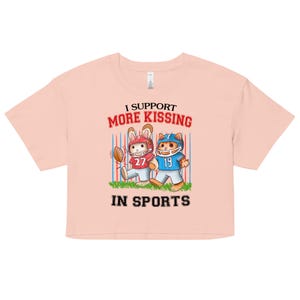 May include: A light pink crop top with the text "I Support More Kissing In Sports." The shirt features a cartoon illustration of a rabbit and a cat in football uniforms. The rabbit wears a red helmet and the cat wears a blue helmet.