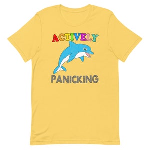 May include: Mustard yellow t-shirt featuring a cartoon dolphin. The dolphin is blue and white, with the words "ACTIVELY" in rainbow colours above it and "PANICKING" below it in black.