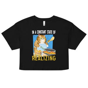 May include: Black cropped t-shirt featuring a graphic of a cat flipping pancakes. The text "In a constant state of realizing" is printed on the shirt.