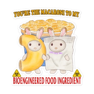 Op de afbeelding: Een sticker met een cartoonillustratie van twee konijnen die verkleed zijn als macaroni en kaas. De tekst "You're the macaroni to my" staat bovenaan de sticker. De tekst "Bioengineered Food Ingredient" staat onderaan de sticker met een biohazardsymbool aan beide kanten.