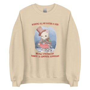 May include: A beige sweatshirt featuring a cartoon illustration of a cat wearing a top hat and holding a red scroll. The text "Wishing all my haters a very being visited by three (3) ghosts tonight" is printed in red on the sweatshirt.