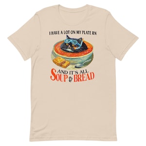 Puede incluir: Una camiseta beige con un gato negro con gafas de sol en un tazón de sopa de tomate con dos rebanadas de pan. El texto en la camiseta dice "I have a lot on my plate rn and it's all soup & bread".