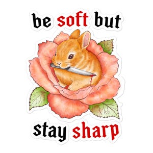 Pode incluir: Um adesivo com um coelho de desenho animado laranja segurando uma faca na boca, aninhado em uma rosa rosa. O texto "be soft but stay sharp" está acima e abaixo da imagem em preto e vermelho.