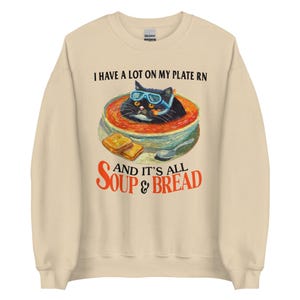 May include: A beige sweatshirt with a black cat wearing sunglasses in a bowl of tomato soup. The text on the sweatshirt reads "I have a lot on my plate rn and it's all soup & bread".