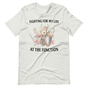 Pode incluir: Uma camiseta branca com fundo cinza mesclado que apresenta um coelho de desenho animado vestindo armadura e segurando uma espada. O texto "FIGHTING FOR MY LIFE AT THE FUNCTION" está impresso na camiseta.