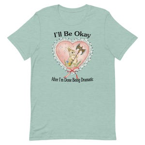 以下が含まれることがあります： 薄い青色のTシャツに、ピンクのハートのデザインが施されています。ハートの中には、小さな斧を持った白いウサギが描かれています。ハートの下には、「I'll Be Okay After I'm Done Being Dramatic」というテキストが印刷されています。
