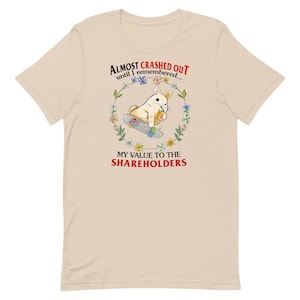 May include: Beige t-shirt with a cartoon rabbit on a skateboard, encircled by a floral wreath. The text says "Almost Crashed Out until I remembered... MY VALUE TO THE SHAREHOLDERS" in black and red.