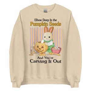 May include: A tan sweatshirt with a cartoon bunny carving a pumpkin. The text reads "Elbow Deep in the Pumpkin Seeds And You're Carving It Out".