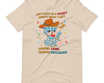 May include: A beige t-shirt with a cartoon blue and white checkered cowboy bear wearing a brown cowboy hat. The text on the shirt reads "Just cause I'm a cowboy doesn't mean I can't conquer anger through gentleness".