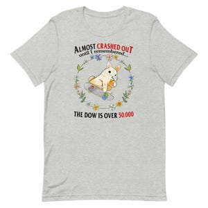 May include: A light gray t-shirt featuring a cartoon rabbit on a skateboard, holding a snack. The design includes text that reads "Almost Crashed Out until I remembered..." and "The Dow is over 50,000."