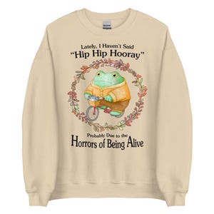May include: A beige sweatshirt with a green frog riding a bicycle in a floral wreath. The text "Lately, I Haven't Said 'Hip Hip Hooray'" and "Probably Due to the Horrors of Being Alive" is printed on the sweatshirt.