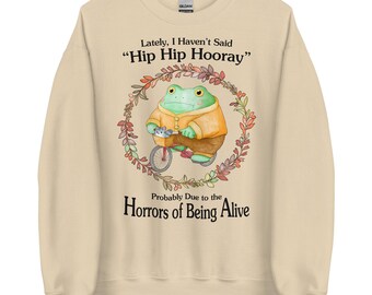 May include: A beige sweatshirt featuring a green frog riding a bicycle within a floral wreath. The text "Lately, I Haven't Said 'Hip Hip Hooray'" and "Probably Due to the Horrors of Being Alive" is printed on the sweatshirt.