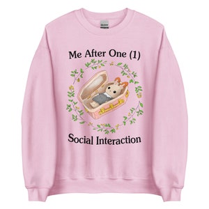Puede incluir: Sudadera rosa claro con el texto "Me After One (1) Social Interaction". El gráfico presenta un conejo de dibujos animados en un ataúd rodeado por una corona floral. La sudadera está hecha de un material suave.