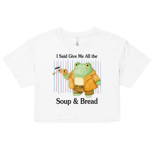 Puede incluir: Top corto blanco con una rana de dibujos animados con una chaqueta marrón y una pistola de juguete. El diseño incluye rayas azules y rojas. El texto en la camiseta dice: "I Said Give Me All the Soup & Bread."