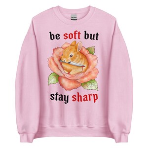 May include: Light pink sweatshirt with a graphic of a brown bunny holding a knife inside a peach-colored rose. The text "be soft but stay sharp" is printed above and below the graphic in black gothic font.