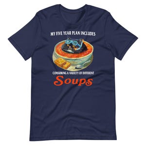 May include: Navy blue t-shirt with a graphic of a black cat wearing goggles in a bowl of soup. The text reads "MY FIVE YEAR PLAN INCLUDES CONSUMING A VARIETY OF DIFFERENT Soups."