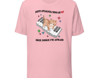 May include: Light pink t-shirt featuring a cartoon kitten playing a piano keyboard. The text "DESPITE APPEARANCES, THINGS ARE NOT" and "OKIE DOKIE I'M AFRAID" are printed above and below the image. Colorful star graphics are also included.