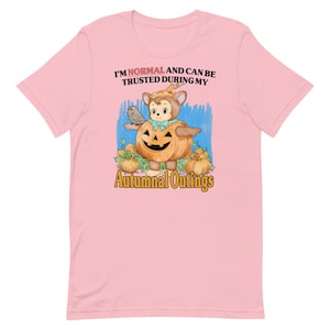 May include: Pink t-shirt featuring a cartoon bear dressed as a pumpkin with the text "I'M NORMAL AND CAN BE TRUSTED DURING MY Autumnal Outings". The bear is holding a bird and surrounded by pumpkins.