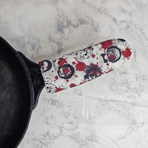 Peut inclure: Une housse de poignée de poêle en fonte noire avec un tissu blanc orné d'un motif répétitif de visages de personnages de films d'horreur et d'éclaboussures de sang rouge. La housse est ajustée sur le manche d'une poêle en fonte noire.