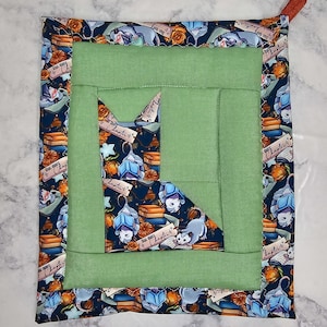 May include: A rectangular fabric potholder featuring a central sage green panel surrounded by a dark blue border patterned with illustrations of opossums reading books, stacks of colourful books, and scrolls labelled One More Chapter.