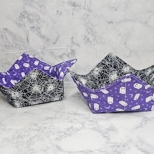 May include: Two fabric bowl cozies featuring a reversible design. One side displays a black background with white spiderwebs, while the other side features a purple background with white ghosts, stars, and crescent moons. Both have four corners.